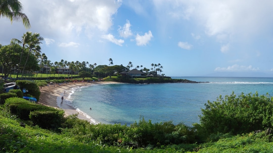 A wide view of a crescent-shaped West Maui bay, sand curving between two green points, with open ocean and a distant island on the horizon